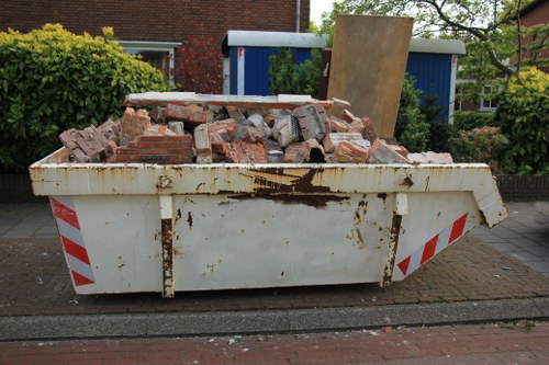A large white metal skip filled with a pile of broken red bricks and concrete debris, located on a paved street in front of a residential area. The skip shows signs of rust and staining, with red and white striped reflective markings on its sides. Behind the skip, there is a blue shed or garage, a wooden panel leaning against it, and greenery including shrubs and trees. The surrounding environment suggests a suburban neighbourhood, and the scene appears to be part of a rubbish removal service in Neasden, London, as indicated by the context of officeclearanceneasden.co.uk and the local postcode inferred from the webpage.