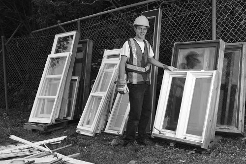 A male worker dressed in a high-visibility safety vest and white hard hat is standing outdoors on a grassy area, holding a white window frame with glass panels. Behind him, several other window frames with various finishes and textures are stacked upright against a chain-link fence, which forms the background of the scene. The window frames are made of wood or uPVC, exhibiting smooth surfaces and neutral tones, with some frames leaning slightly or stacked in front of others. The environment appears organized and indicative of a rubbish removal or clearance activity typically associated with property or office clearance services in Neasden. The ground shows part of a driveway or pavement, with additional loose window parts lying in front, suggesting ongoing clearance or loading activities. The natural lighting highlights the textures of the glass and frame materials, creating a professional and factual snapshot relevant to rubbish removal services on the [POSTCODE] area.