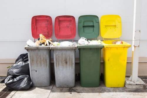 A row of four large wheelie bins positioned outside a building, each with a different coloured lid — red, red, green, and yellow — standing on a concrete surface adjacent to a white wall. The grey bins in the foreground are filled with mixed household waste, including paper, plastic bags, and other rubbish, with some waste spilling over the tops. To the left, there are two black garbage bags placed against the grey bins, and the yellow bin has a small cardboard box and some loose debris visible on its open lid. The green bin is partially obscured behind the grey bins, with its lid slightly open. The environment suggests an area designated for waste disposal and rubbish removal services, consistent with the types of containers used for general waste collection in urban settings like Neasden. The lighting is natural, and the scene captures a typical waste storage setup in a residential or commercial neighbourhood within northwest London.
