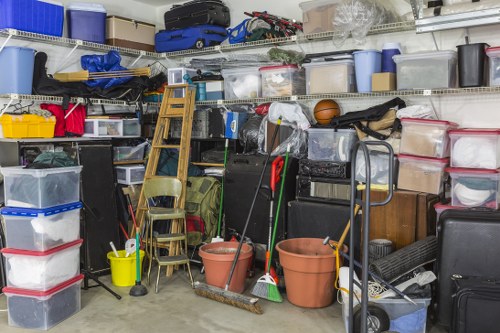 A cluttered storage space or garage filled with various items including transparent plastic storage boxes stacked on shelves, some with red or black lids, and others open or partially open revealing contents like supplies and tools. There are two large terracotta flower pots leaning against a shelving unit that holds miscellaneous objects such as sports equipment, cardboard boxes, and plastic containers. A yellow mop bucket with a mop inside sits on the concrete floor next to two wooden chairs stacked one behind the other. Several cleaning tools, including a broom and a dustpan, are visible among the clutter. A black trolley or hand truck is positioned on the right side, near additional storage boxes and a dark suitcase or equipment case. The background shows a metal wire shelving system holding items indicative of an organised but heavily used storage area, suggesting it could be part of a rubbish clearance or decluttering project typical of Office Clearance Neasden services. The environment appears well-lit, possibly by overhead fluorescent lighting, with a mix of different textures such as plastic, wood, and metal in the stored objects.