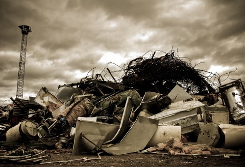 A large pile of assorted discarded debris and scrap materials is shown in an outdoor setting, with a cloudy sky overhead. The waste includes broken appliances, metal fragments, plastic parts, and tangled ropes, all piled haphazardly on the ground. Some items are partially covered in rust and dirt, indicating exposure to the elements. In the background, there is a tall, slender industrial tower or antenna on the left side, with no other structures visible nearby. The scene appears to be in a waste or recycling collection area, possibly in or near Neasden, London, serving rubbish removal services. The overall atmosphere is overcast, with diffused lighting casting soft shadows across the jumble of waste, emphasizing the textures of metal, plastic, and organic materials in the pile. This detailed visual reflects the type of clutter managed by office clearance and rubbish removal specialists like Office Clearance Neasden.