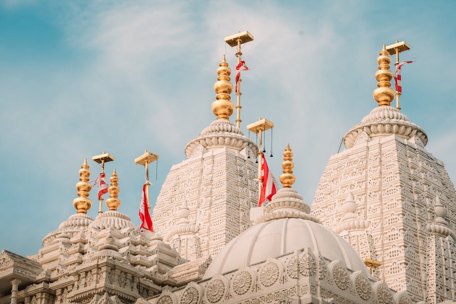The image depicts a large outdoor pond with a central fountain spraying multiple streams of water into the air, situated in front of a grand Hindu temple complex with intricate white stone carvings an