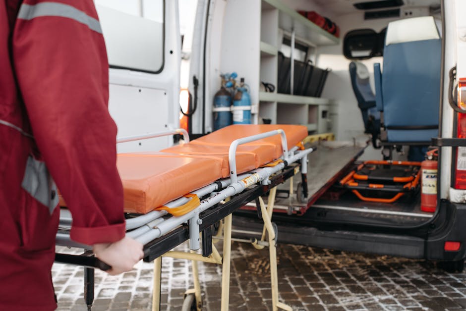 The image depicts a healthcare professional, wearing a red uniform, loading an orange emergency stretcher with padded cushioning onto a wheeled trolley at the rear of a white emergency vehicle, likely