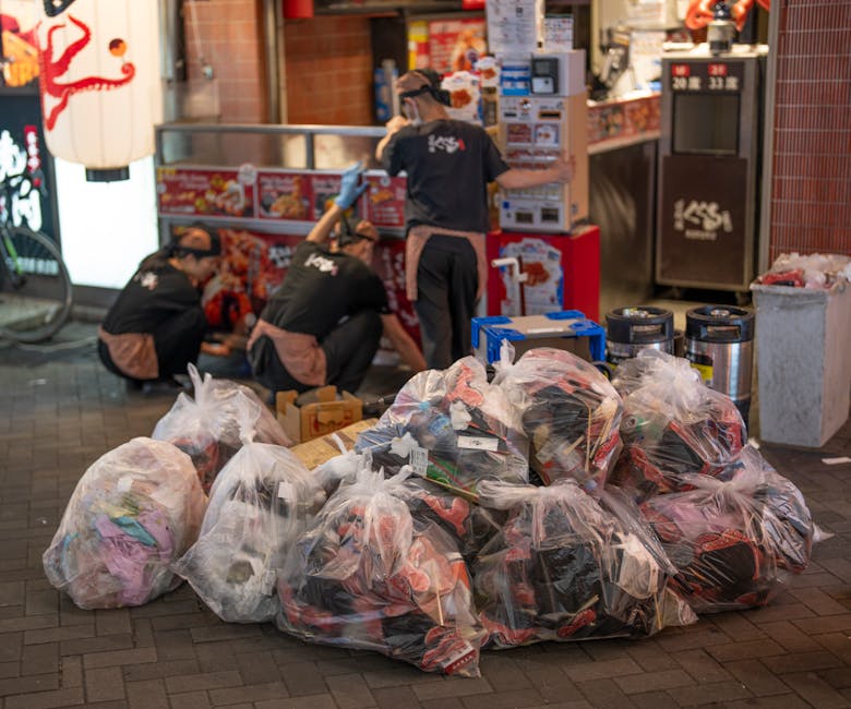Two waste management workers in high-visibility green and orange uniforms are seen operating a large industrial rubbish compactor on a city street. One worker is standing on the ground, with his back