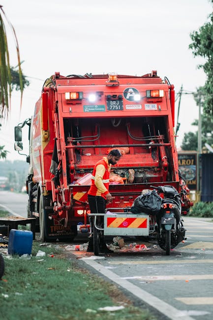 A large pile of mixed waste and rubbish, including cardboard boxes, plastic bags, and paper waste, is overflowing from open recycling bins situated on a paved area outside a commercial building. The b